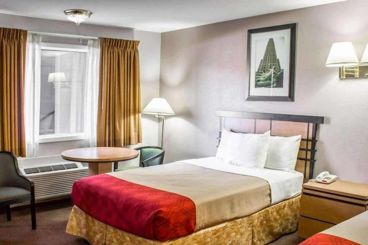 A hotel room with a bed, a desk, a chair, a window with curtains, a lamp, and a framed picture on the wall.