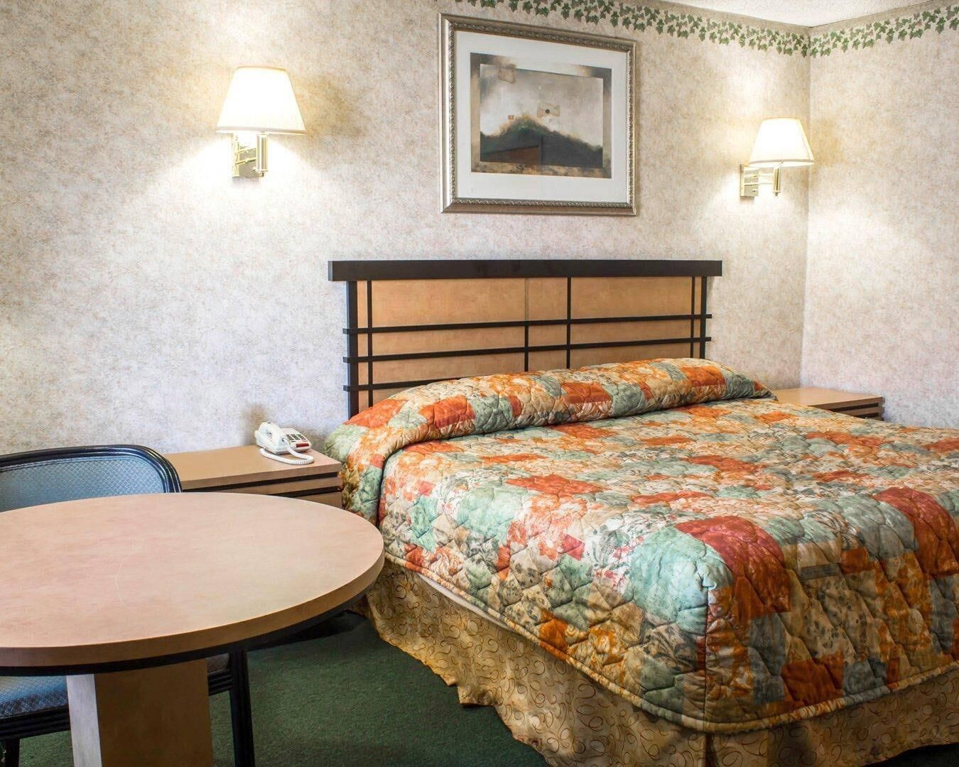 A hotel room with a bed, a round table, two bedside lamps, a framed picture on the wall, and a telephone on the nightstand.