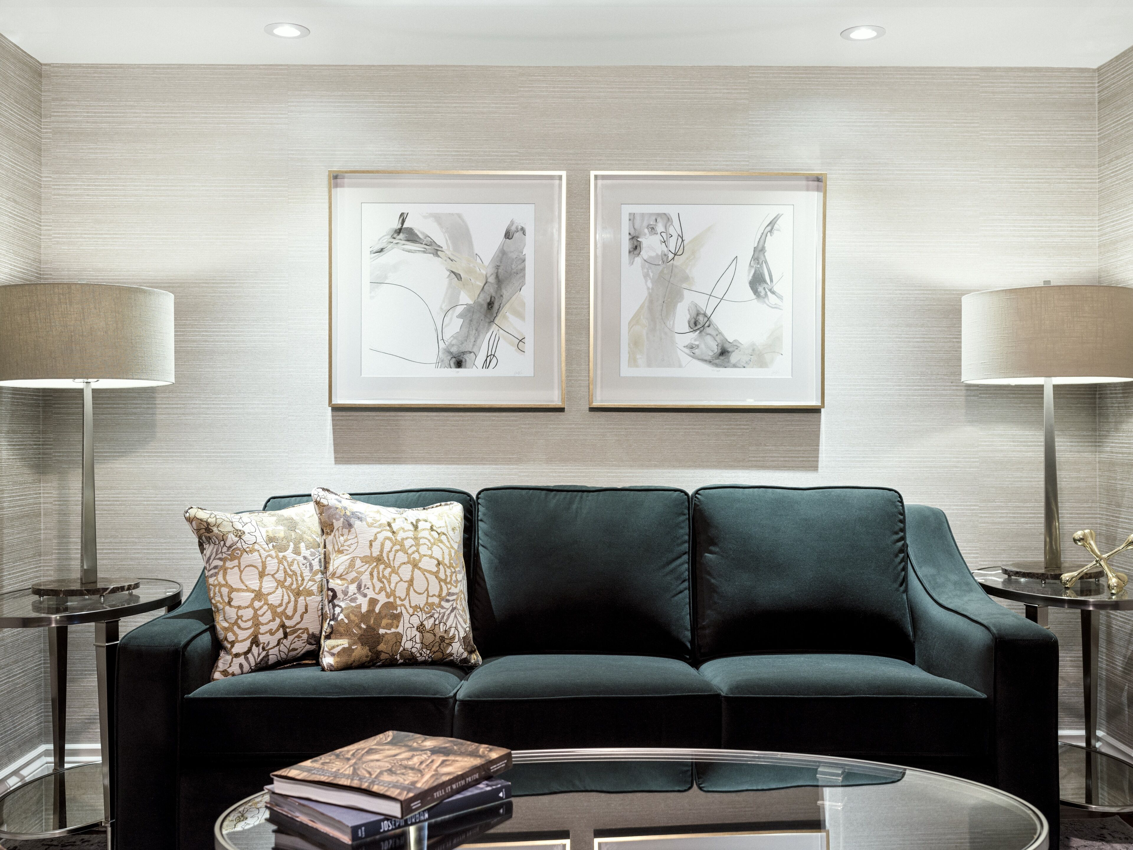 A modern living room with a dark green sofa, two framed abstract paintings, a side table with a lamp, and a coffee table with books.