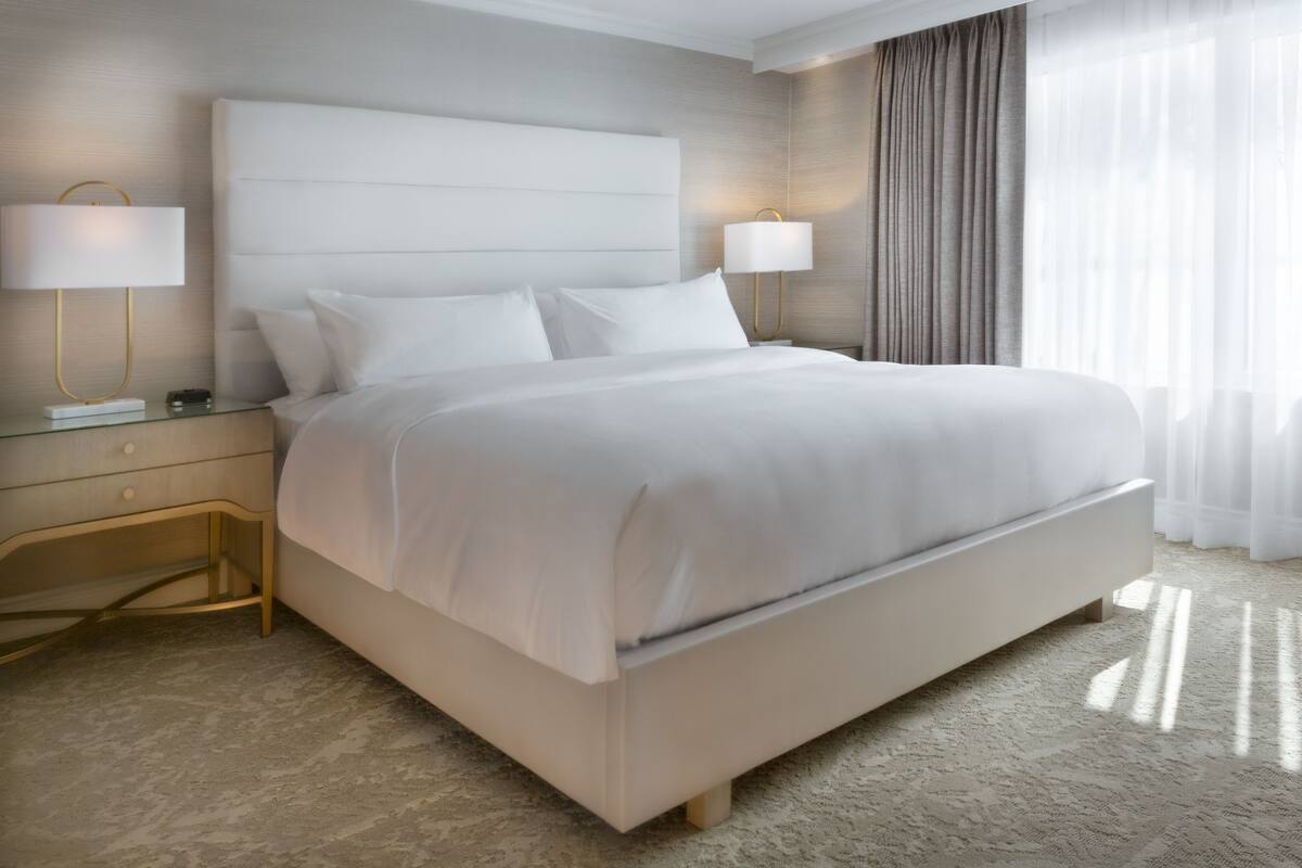 Une chambre d’hôtel moderne avec un grand lit, deux tables de chevet avec des lampes et une vue sur l’extérieur à travers des rideaux légers.