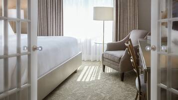 A hotel room with a bed, a chair, a lamp, and a view of the outdoors through a window with curtains.