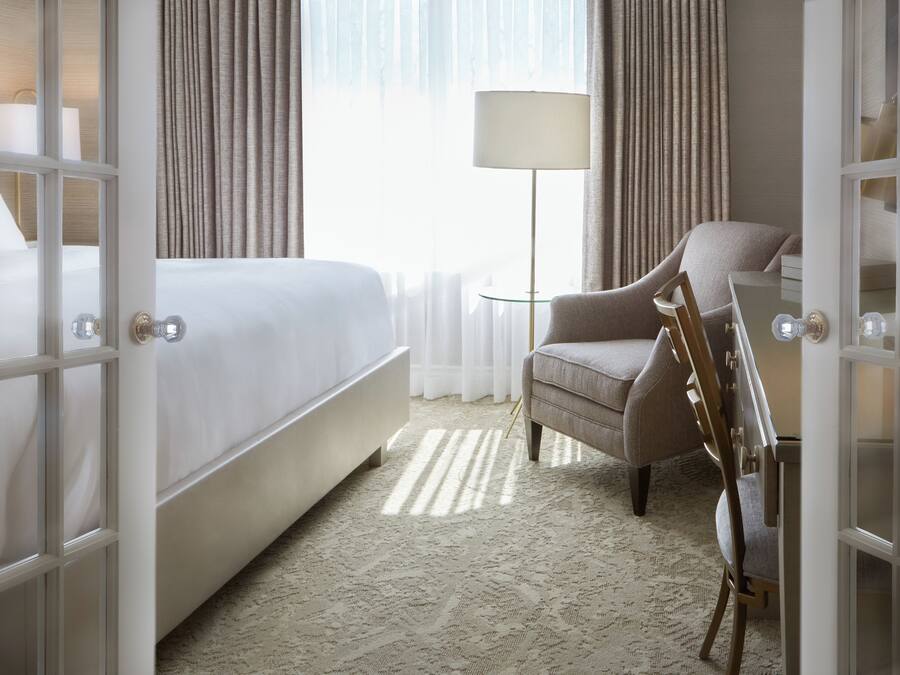 A hotel room with a bed, a chair, a lamp, and a view of the outdoors through a window with curtains.