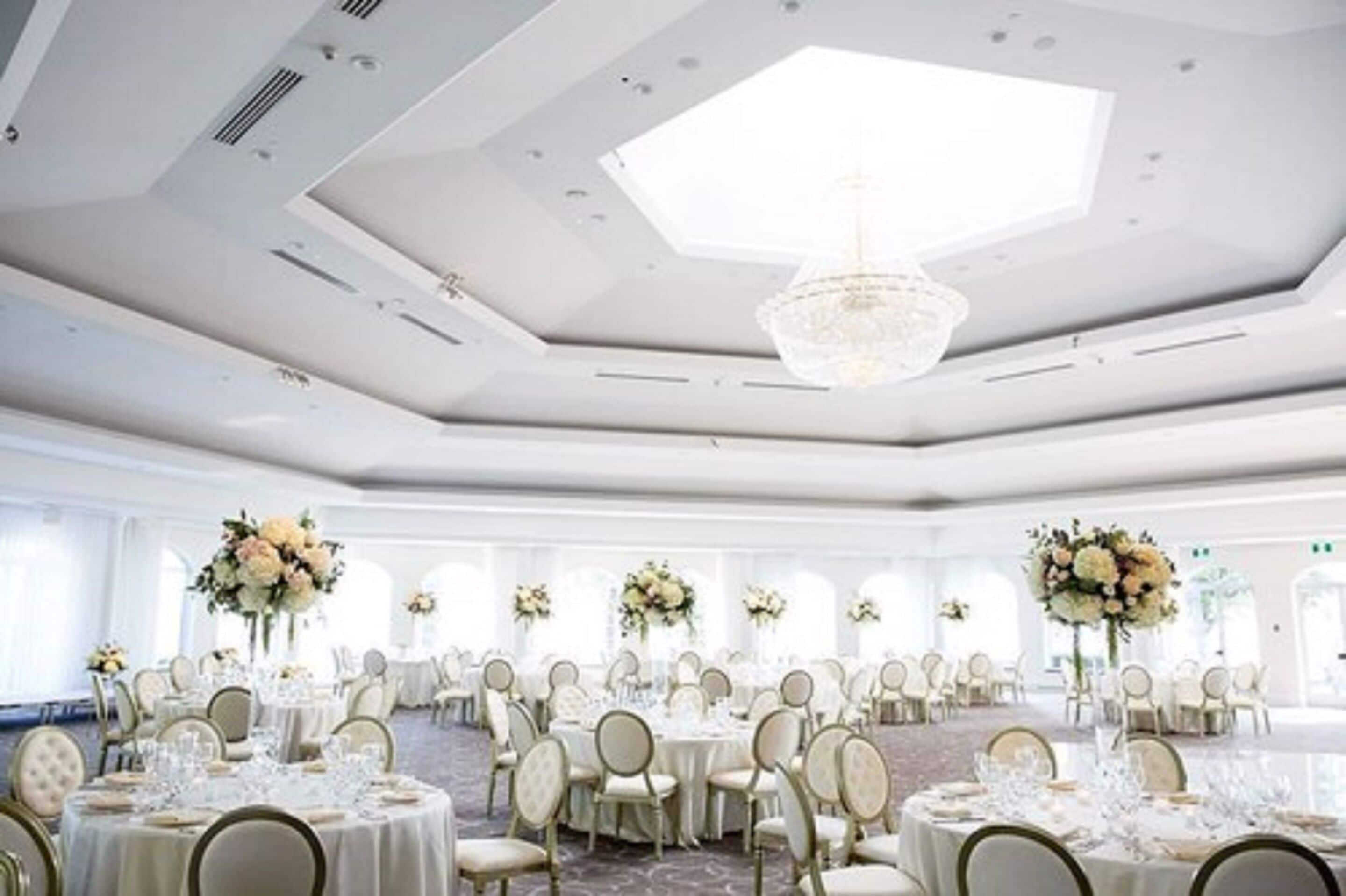 Un amplio salón de banquetes con mesas redondas, sillas blancas y centros de mesa florales.