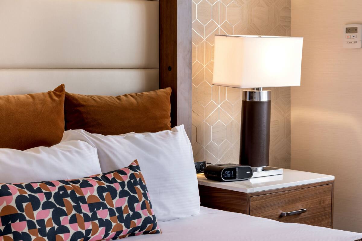 A hotel room with a bed, pillows, a bedside table with a lamp, and a digital clock.