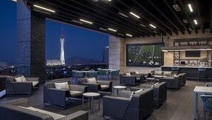 A rooftop bar with wicker furniture, a large TV screen, and a view of a tower.