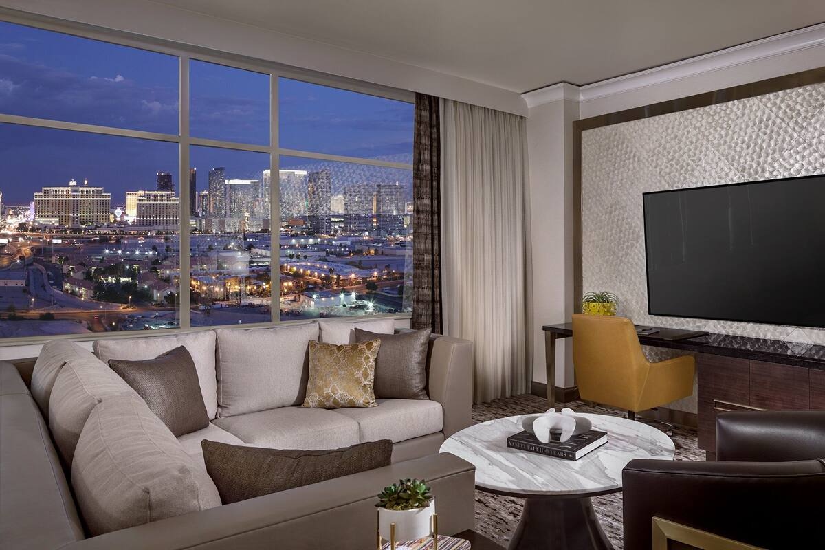 A modern hotel room with a large window offering a city view, a flat-screen TV, a sofa, a coffee table, and a leather armchair.