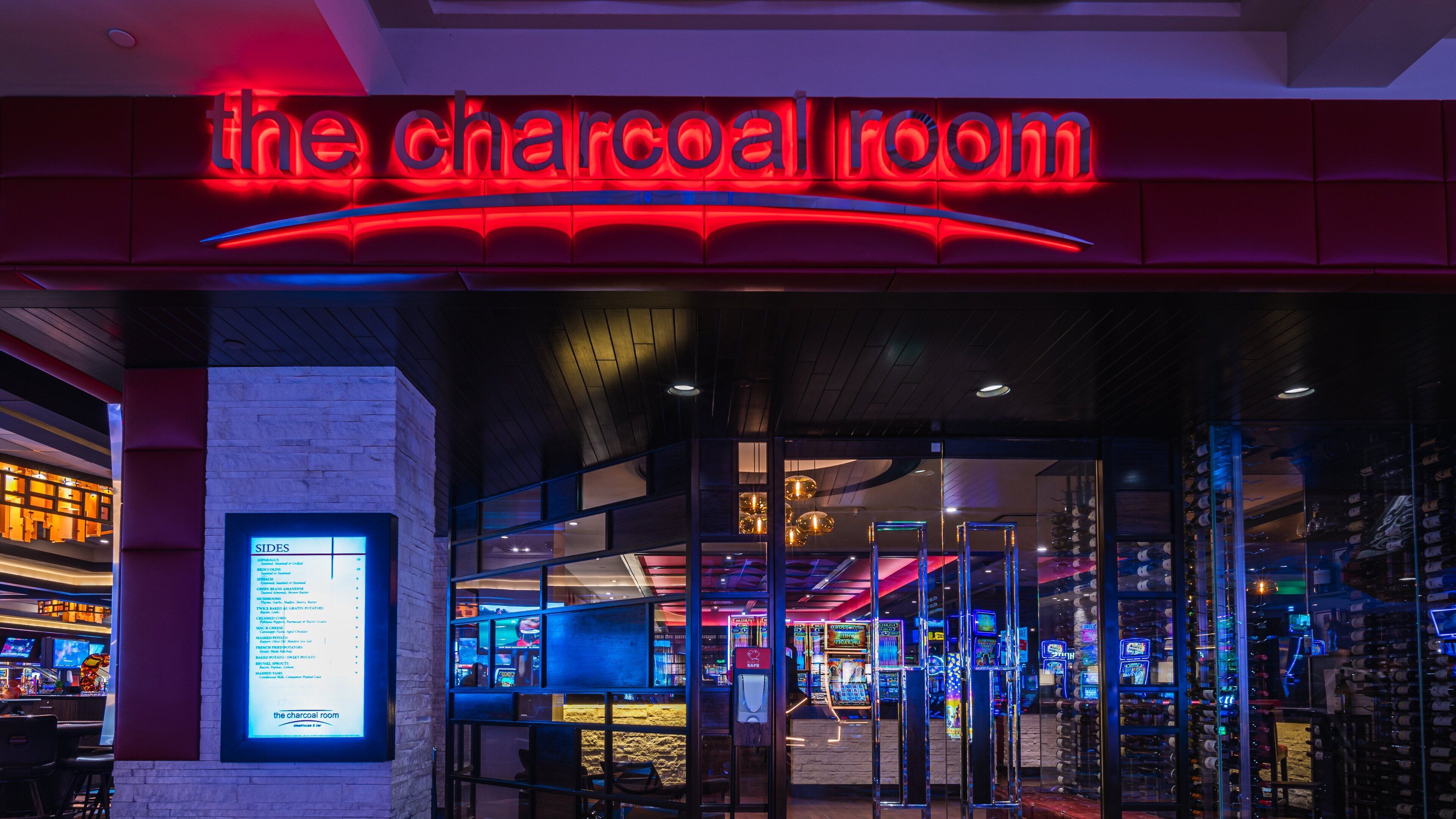 The entrance to The Charcoal Room with a neon sign, glass doors, and a visible menu board listing various food items.