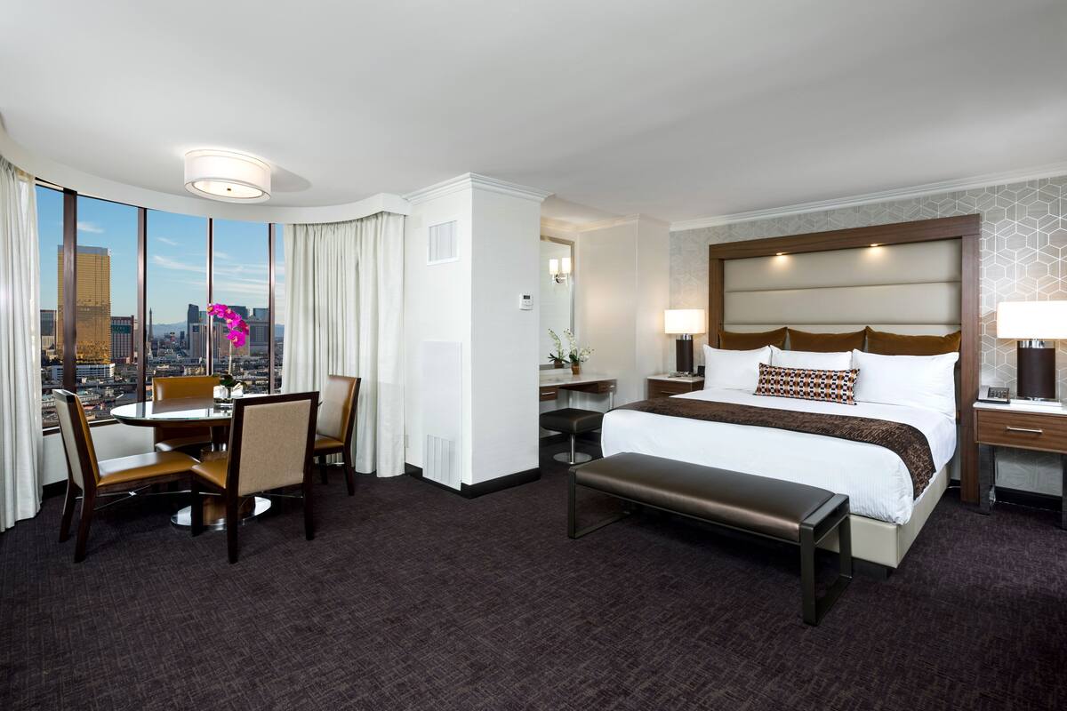 A hotel room with a large bed, a desk, a chair, a bench, and a view of the city.
