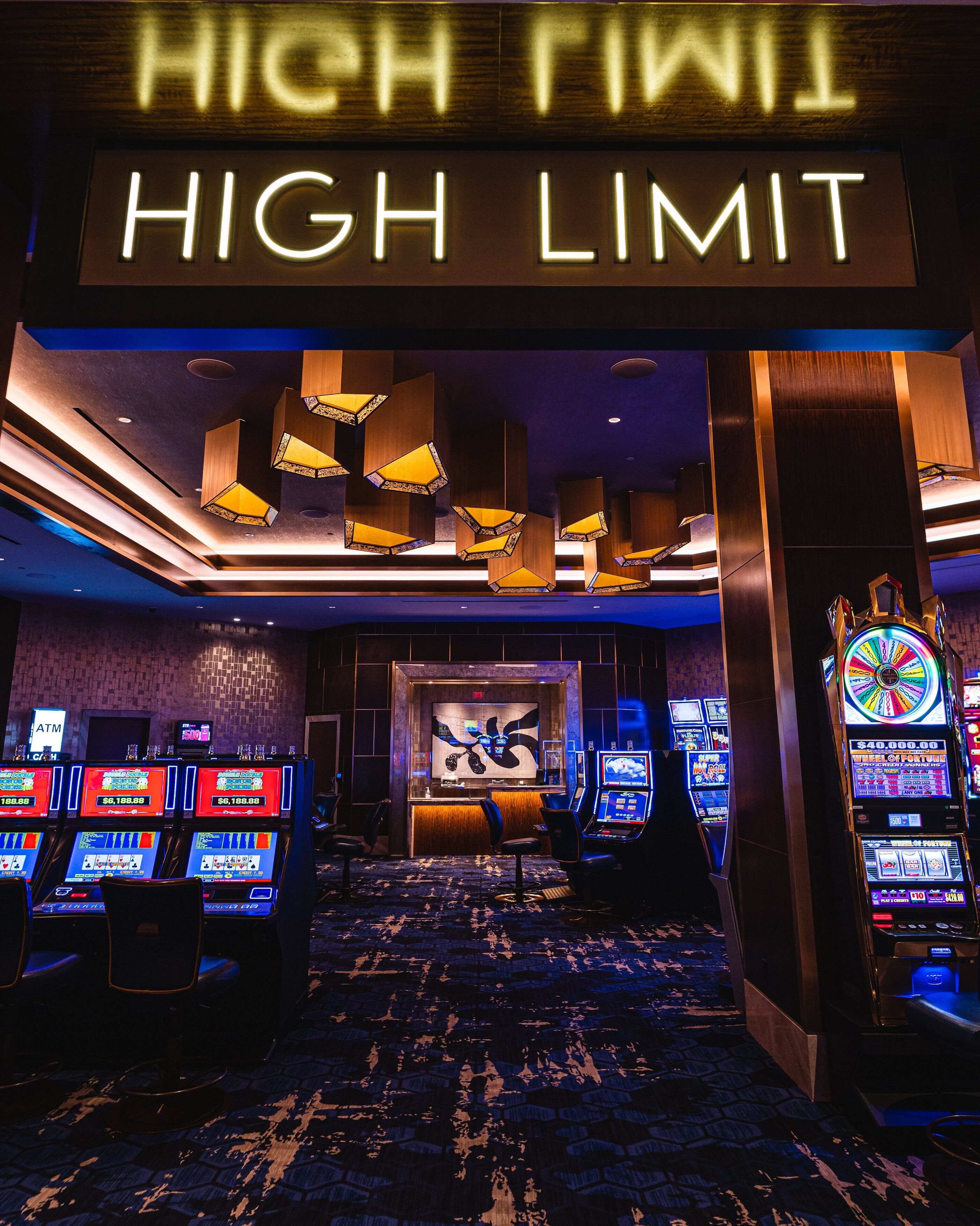 Interior de un casino con máquinas tragamonedas, un letrero de apuestas altas y una gran ruleta.