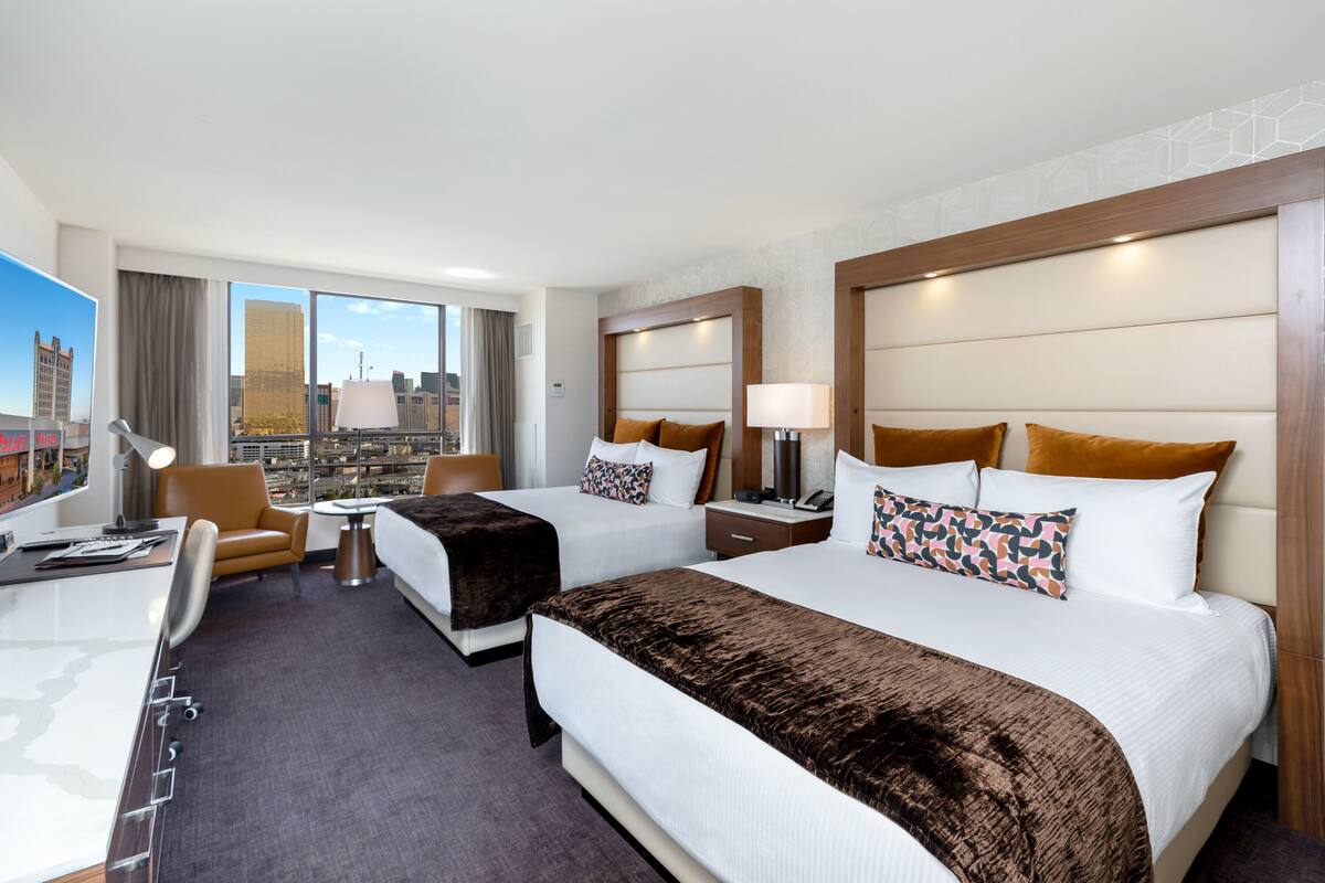 A hotel room with two beds, a desk, a chair, and a view of the cityscape.