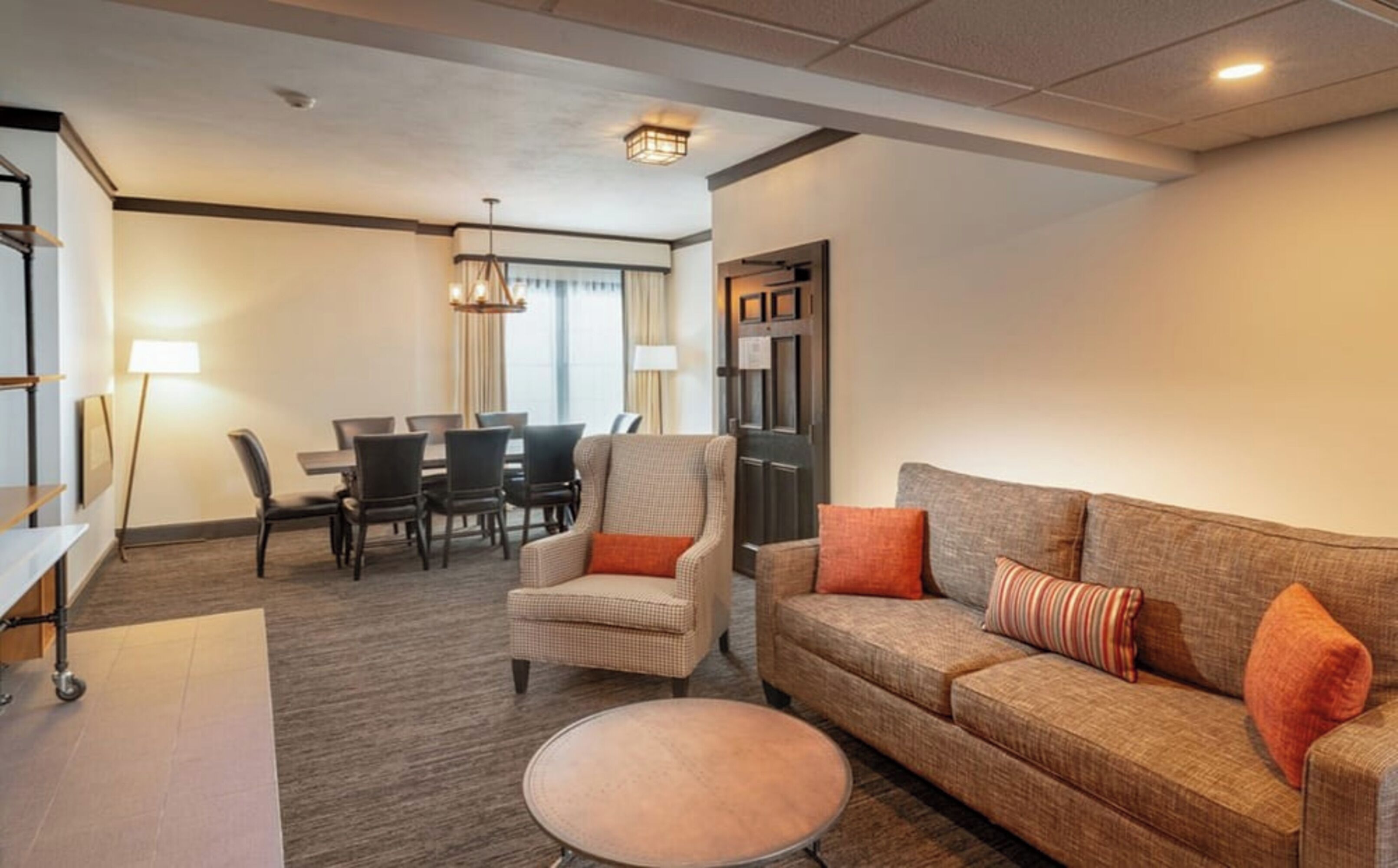 A hotel room with a sofa, armchair, dining table, chairs, and a lamp.