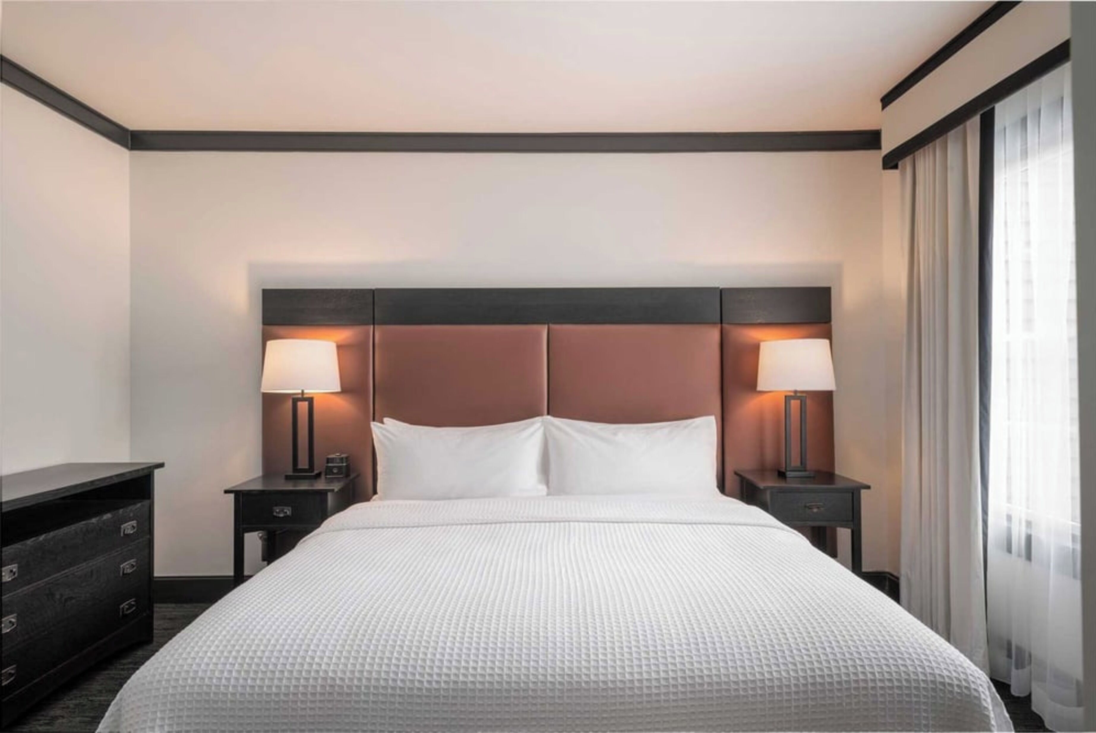 Habitación de hotel con una cama grande, dos mesitas de noche con lámparas, un aparador y una ventana con cortinas.