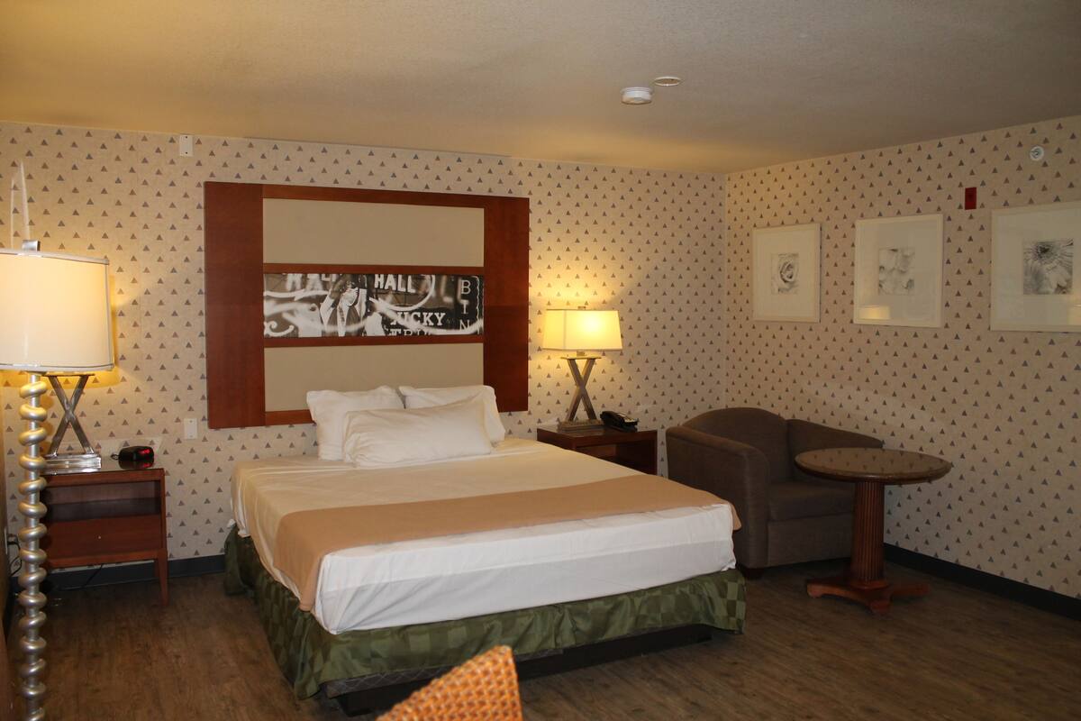 A hotel room with a bed, two bedside tables, a chair, a small table, and a wall with framed pictures.