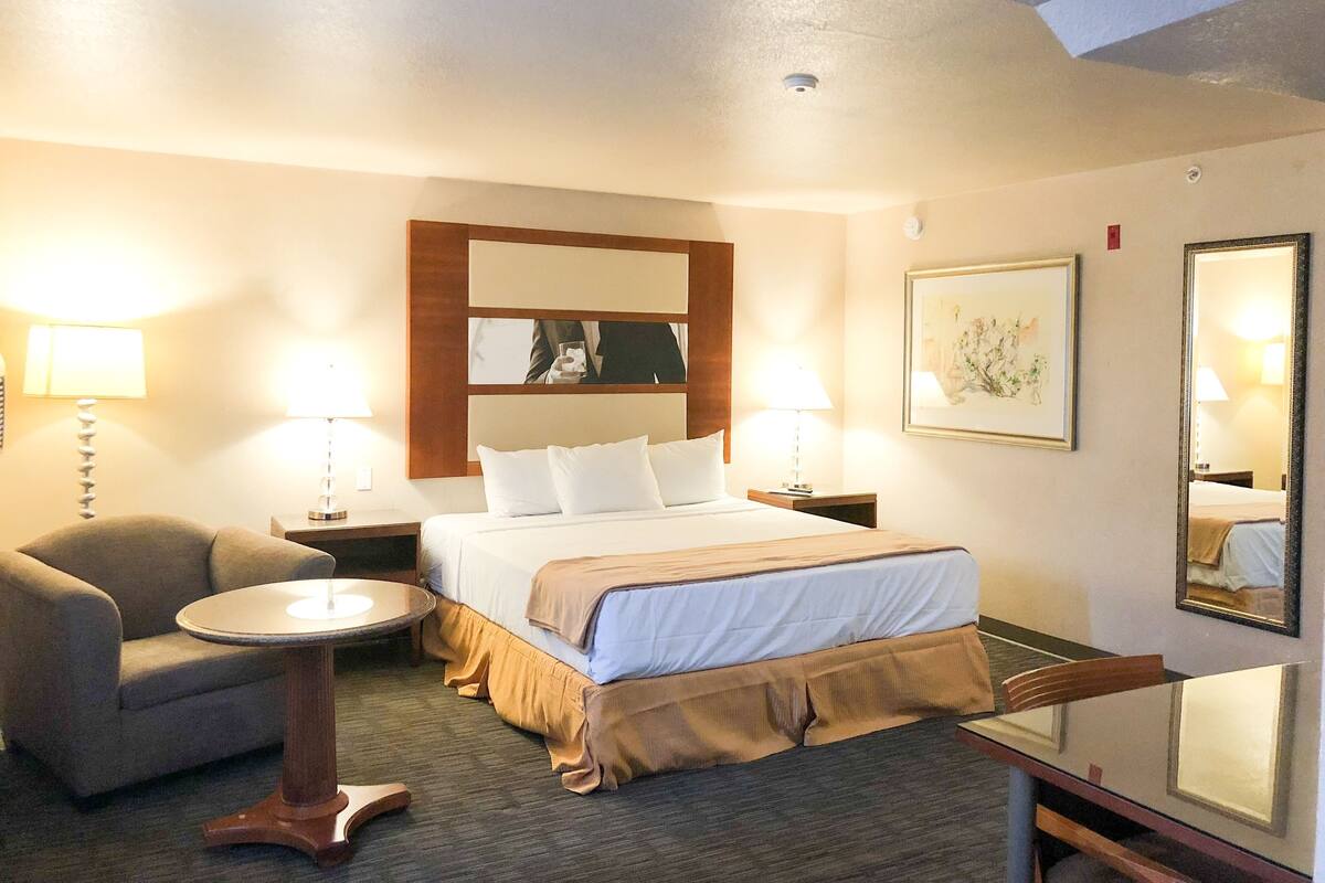 A hotel room with a bed, two bedside tables with lamps, a chair, a small round table, and a mirror.