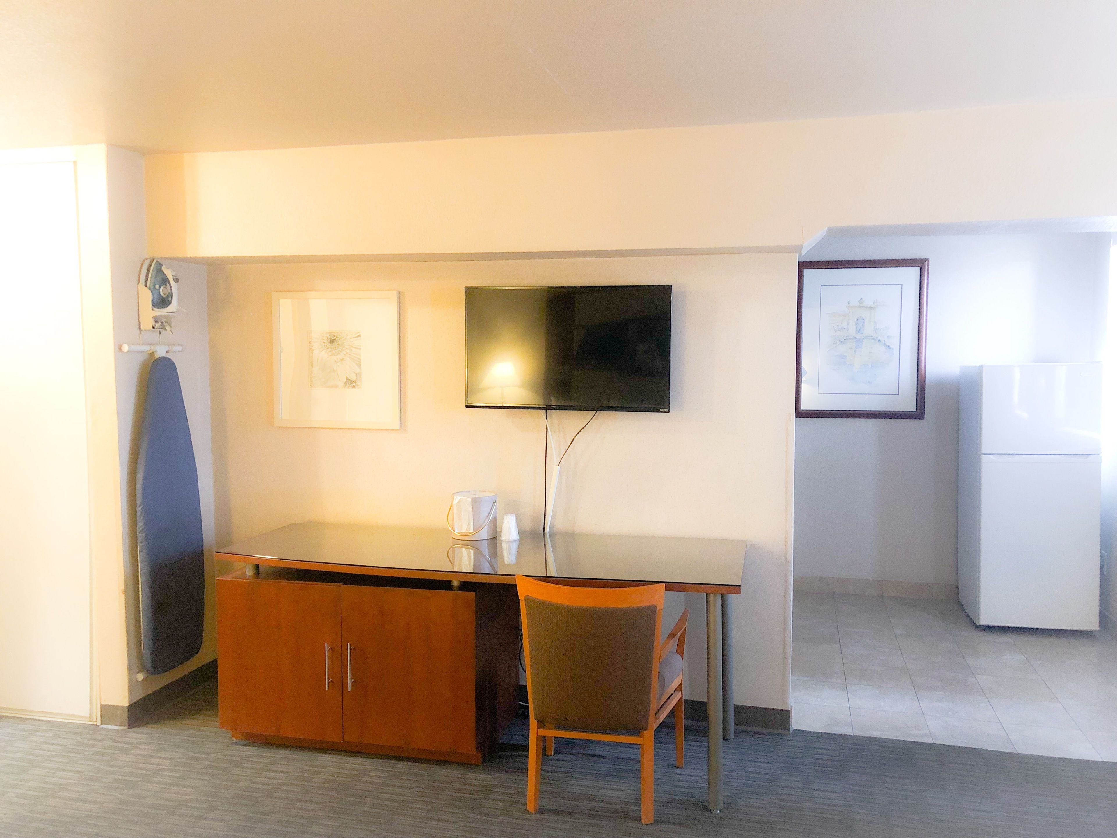 Una habitación pequeña con escritorio, silla, tabla de planchar, televisor y refrigerador.