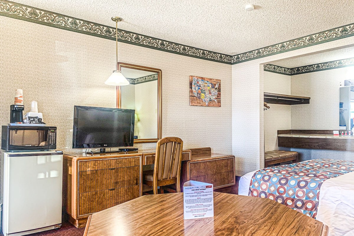 Ein Hotelzimmer mit einem Holztisch, einem Fernseher, einer Mikrowelle, einem Kühlschrank, einem Bett mit gemusterten Bettwaren, einem Spiegel und einer Wand mit dekorativer Bordüre.