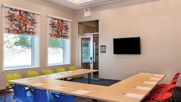 Une salle de conférence avec une longue table, des chaises colorées, un téléviseur fixé au mur et des fenêtres équipées de stores.