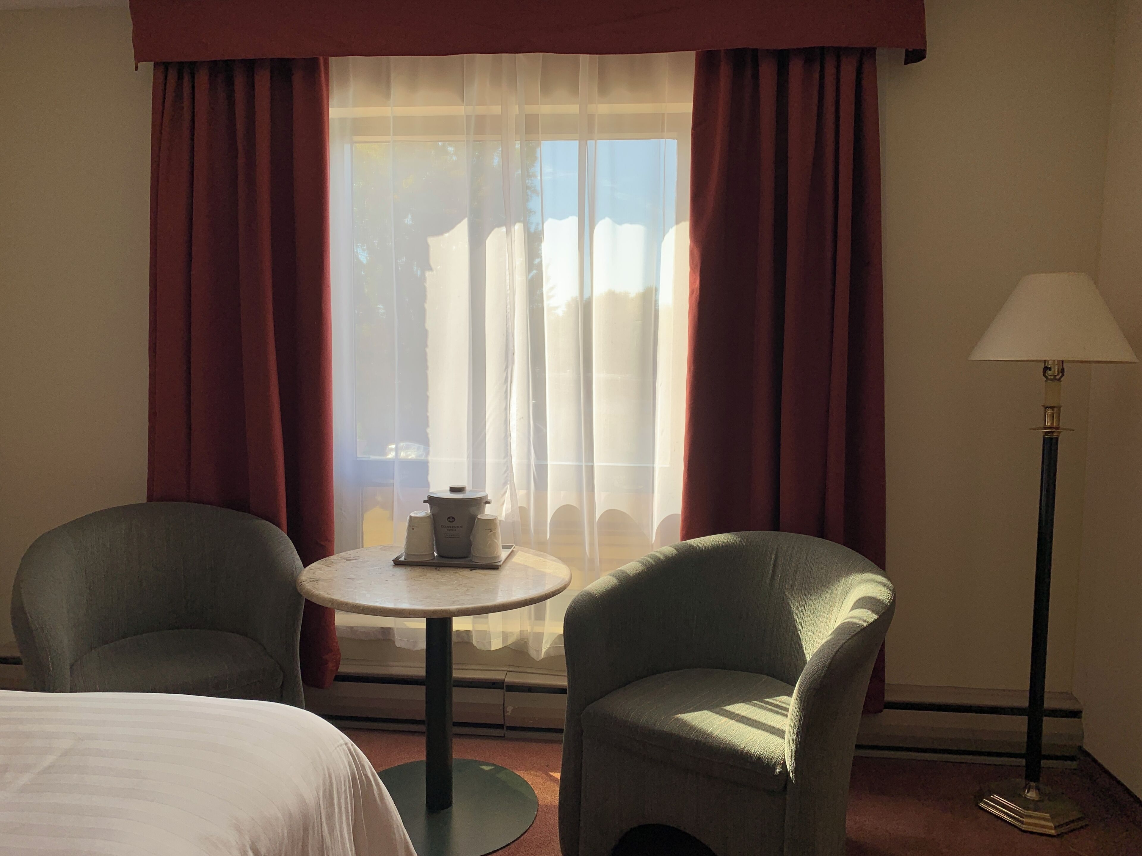 A hotel room with a bed, two armchairs, a small round table with a lamp, and a window with curtains.