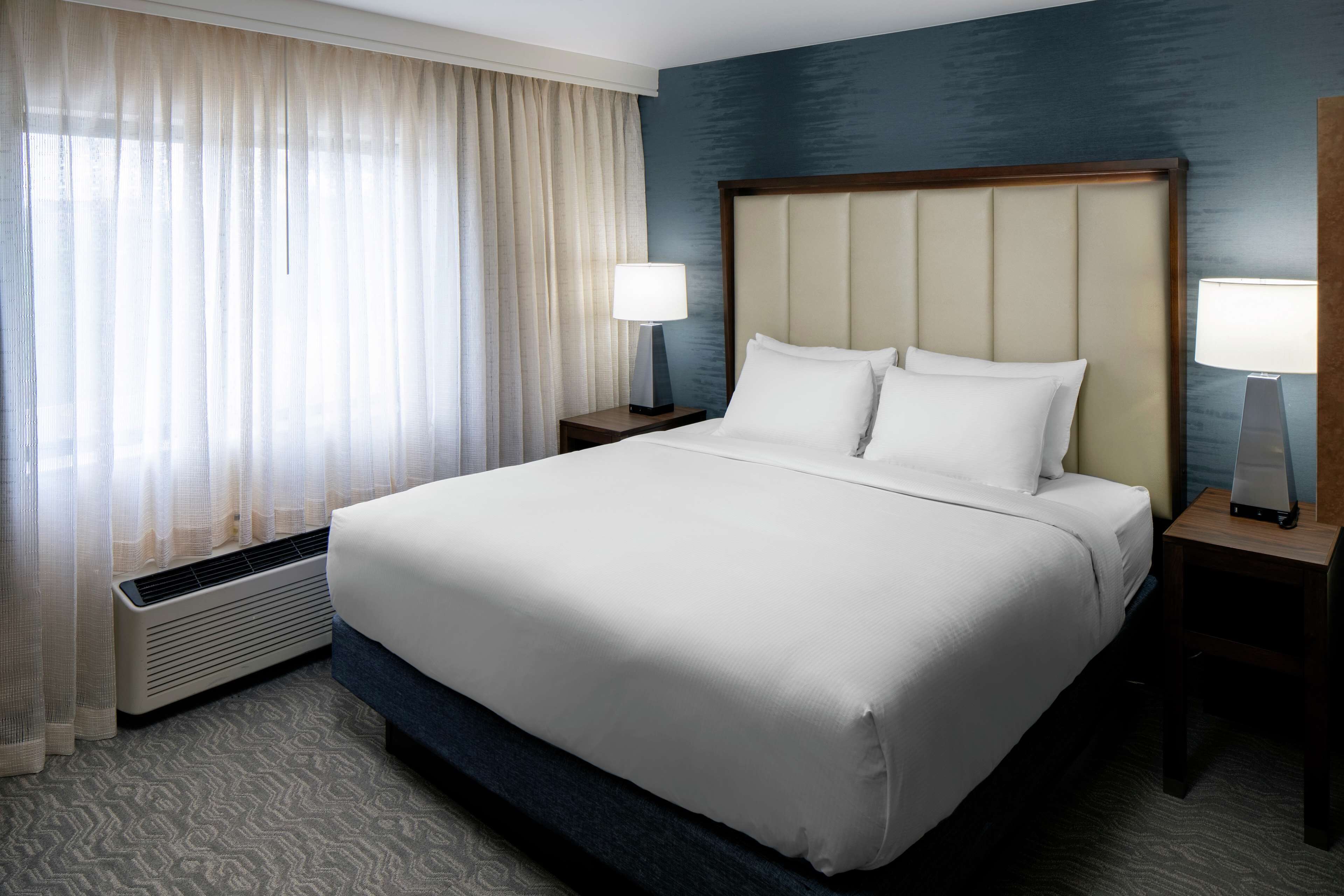 A hotel room with a large bed, two bedside tables with lamps, a window with sheer curtains, and a wall heater.