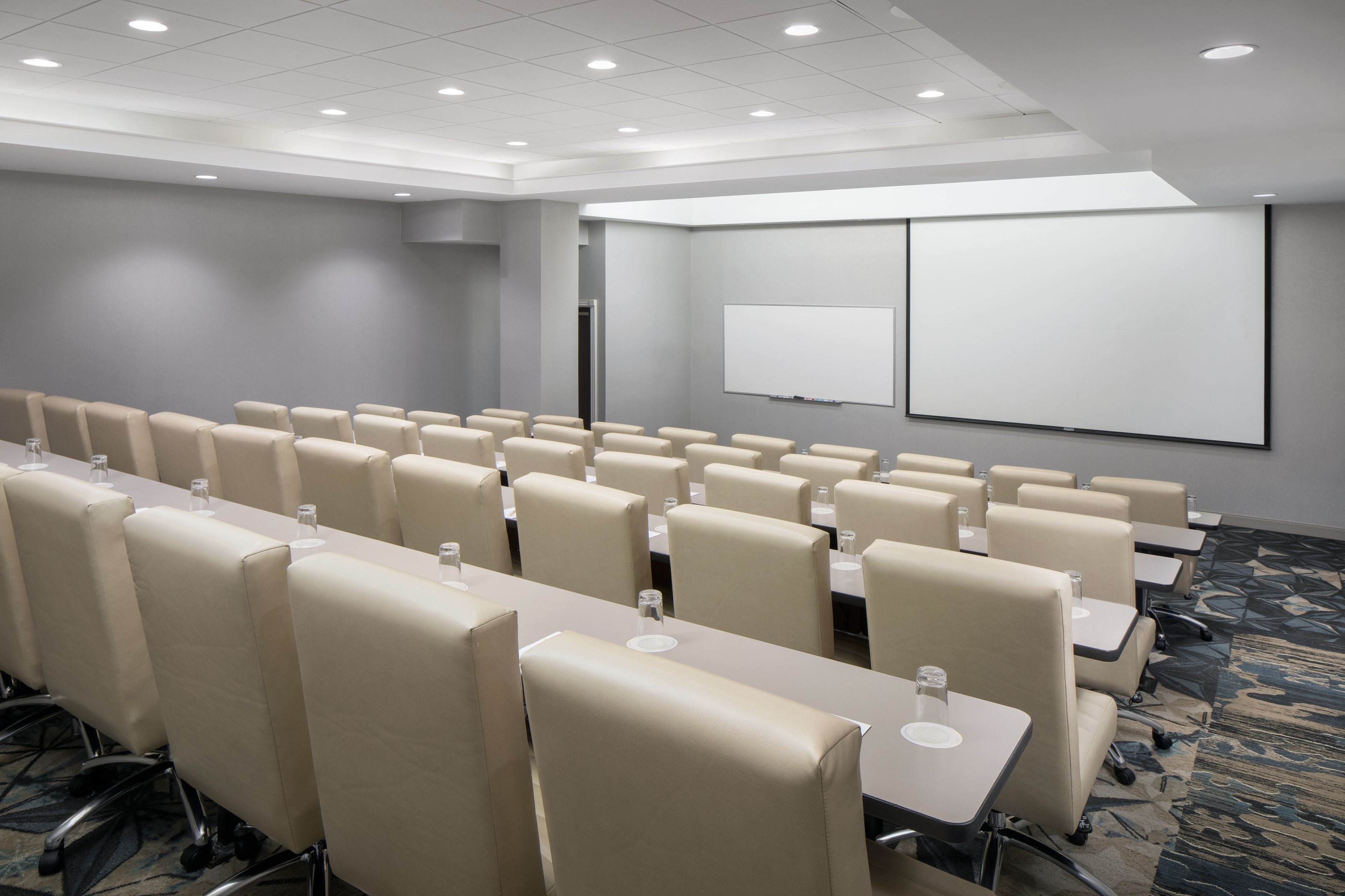 Un salón de conferencias con filas de sillas y mesas color beige, una pantalla de proyección blanca y piso alfombrado.