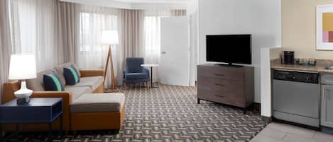 A hotel room with a sofa, armchair, TV, and kitchenette.