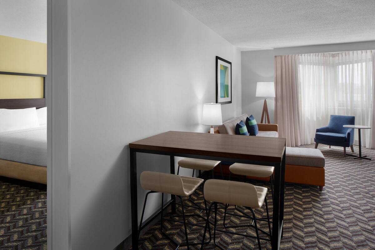 A hotel room with a bed, a desk with chairs, a lamp, a chair, and a window with curtains.