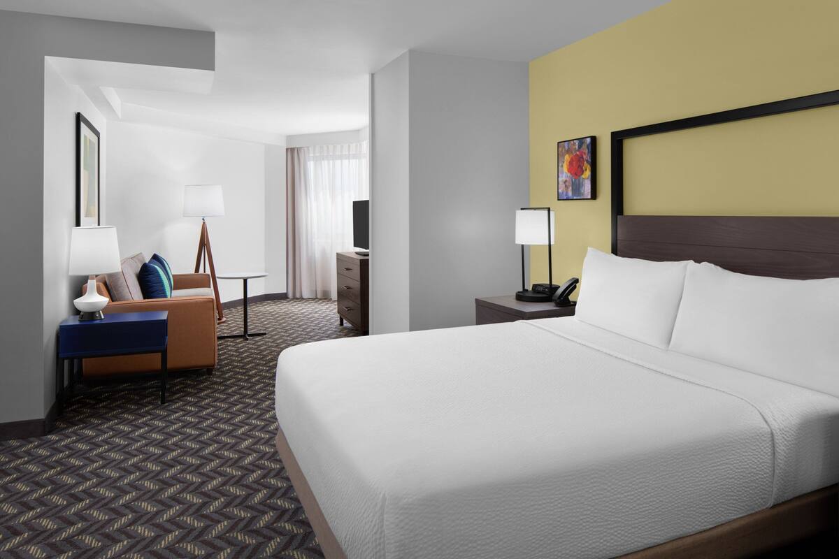A hotel room with a large bed, a desk, a chair, a lamp, and a painting on the wall.