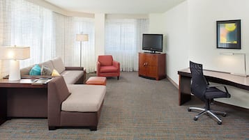 Habitación de hotel con sofá, reposapiés, escritorio, silla, televisión y una obra de arte en la pared.