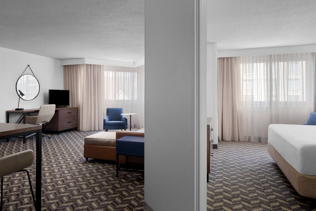 A hotel room with a bed, a desk with a chair, a small table, a sofa, a TV, and curtains.