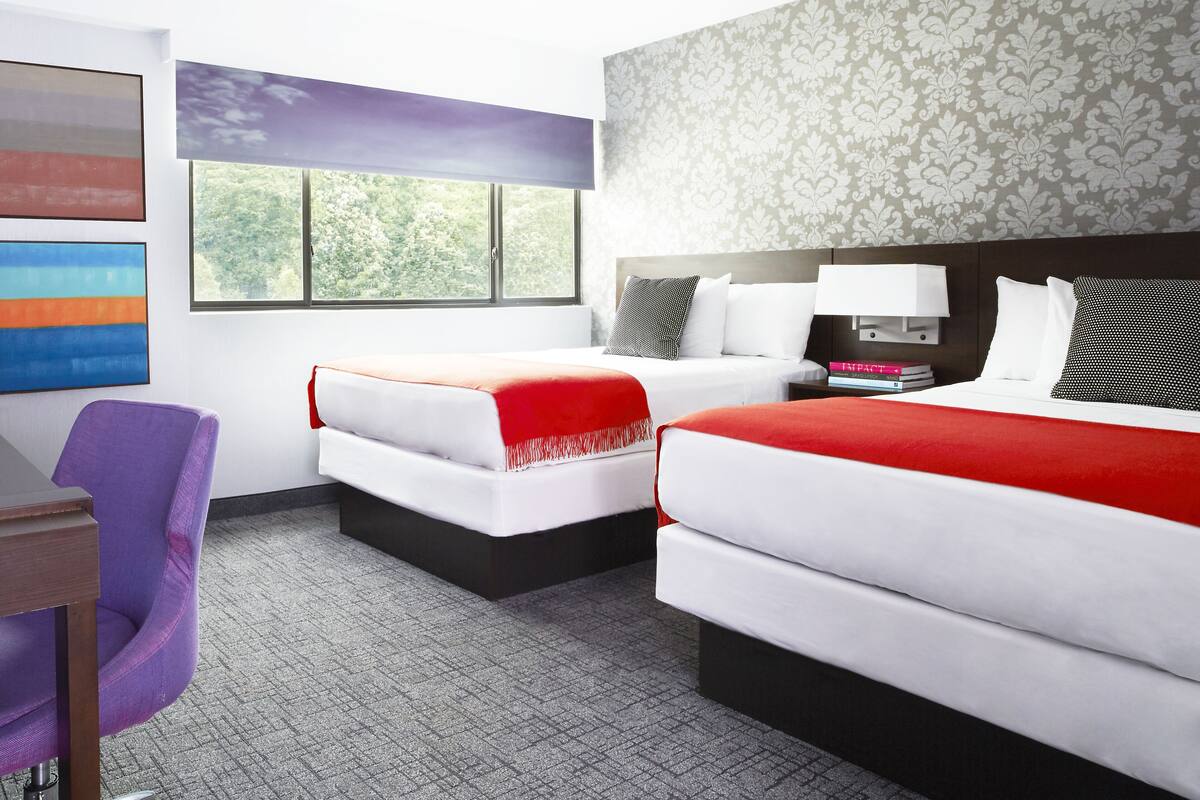 Habitación de hotel con dos camas, un escritorio, una silla morada y una ventana que da a un paisaje de árboles.
