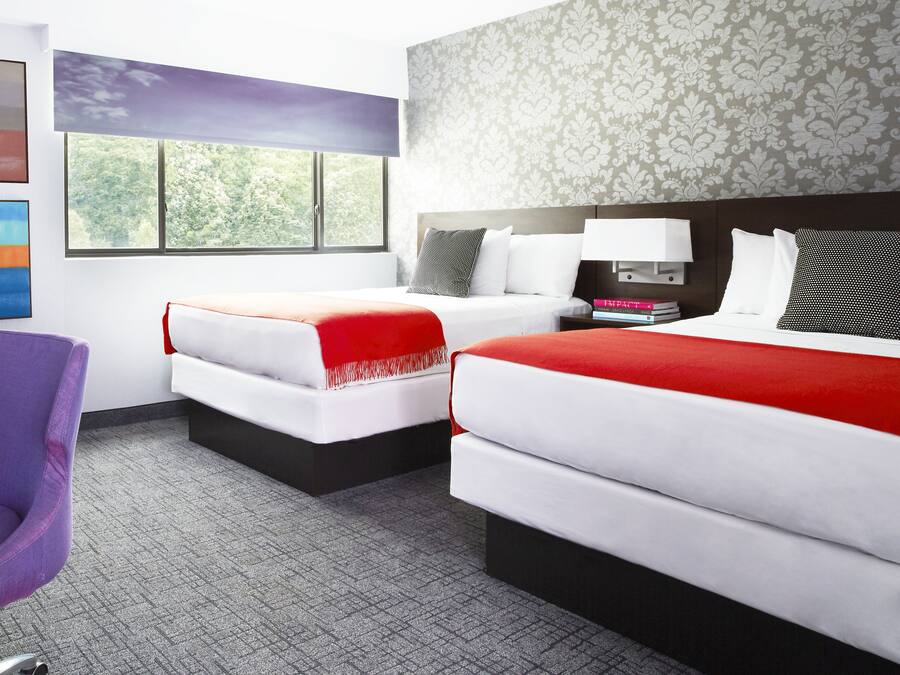 Habitación de hotel con dos camas, un escritorio, una silla morada y una ventana que da a un paisaje de árboles.