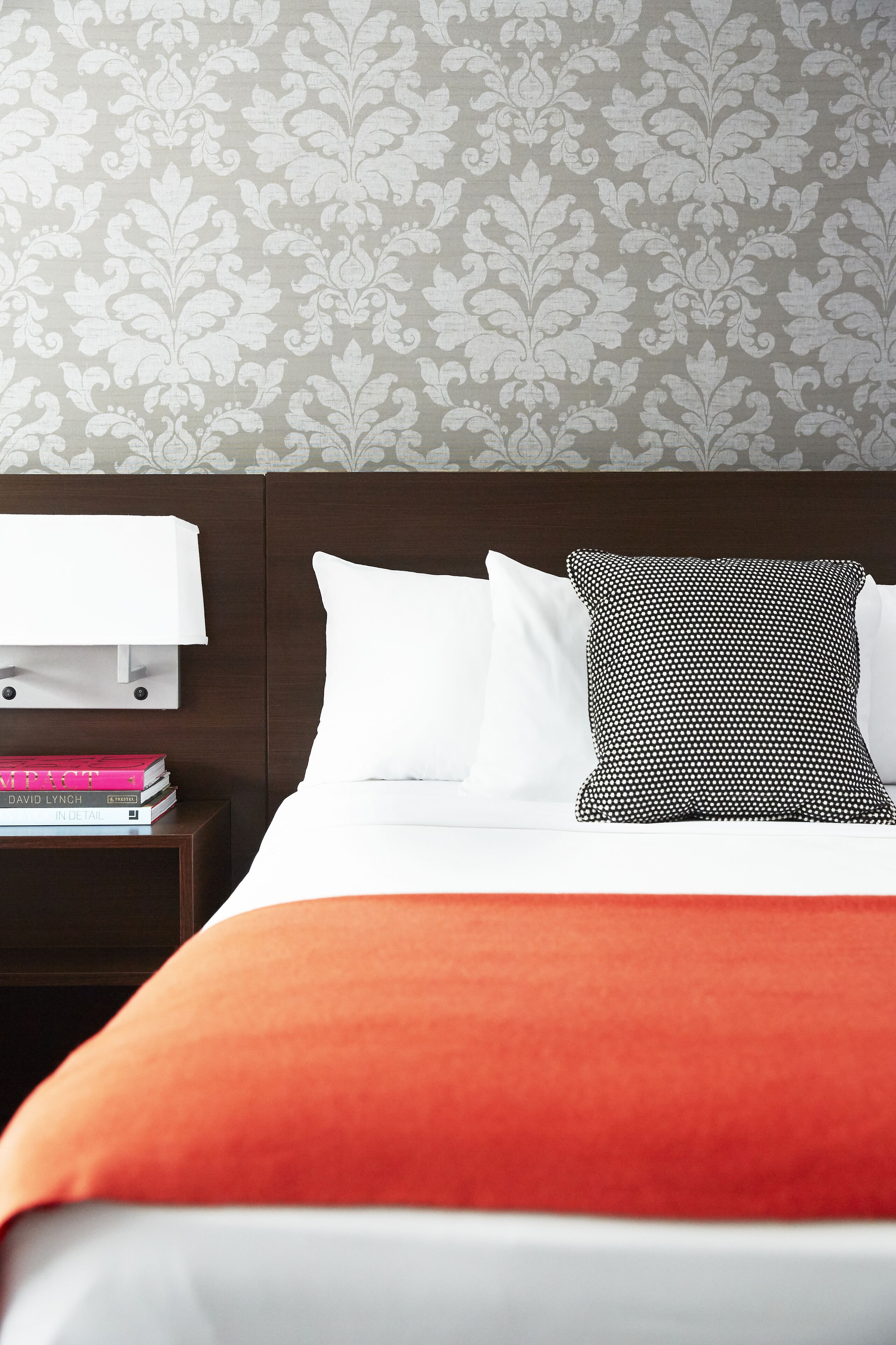 A neatly made bed with white and black pillows, an orange throw blanket, and a nightstand with a white lamp and a few books.