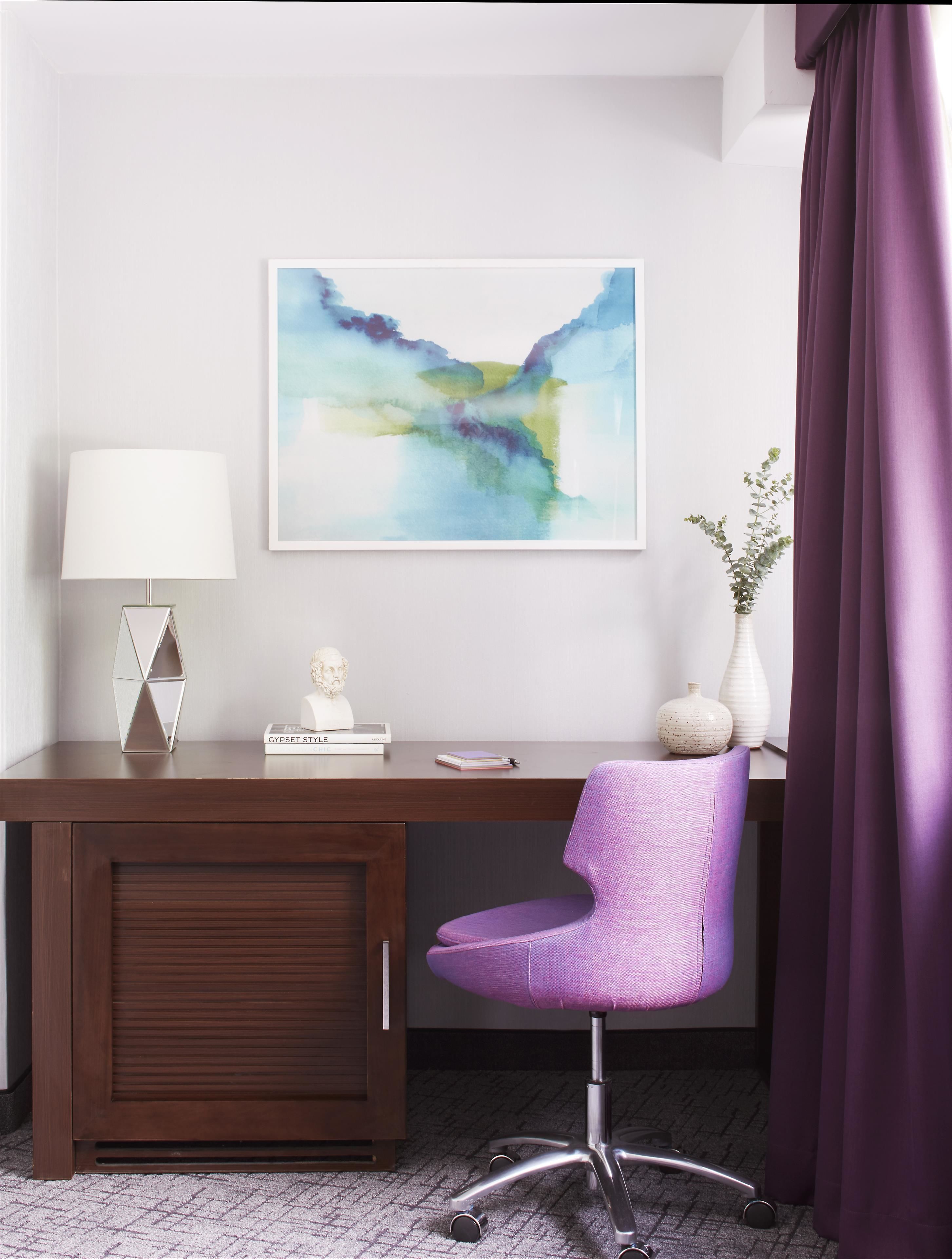 A modern office space with a dark wood desk, a purple office chair, a white lamp, a framed abstract painting, and a vase with a plant.