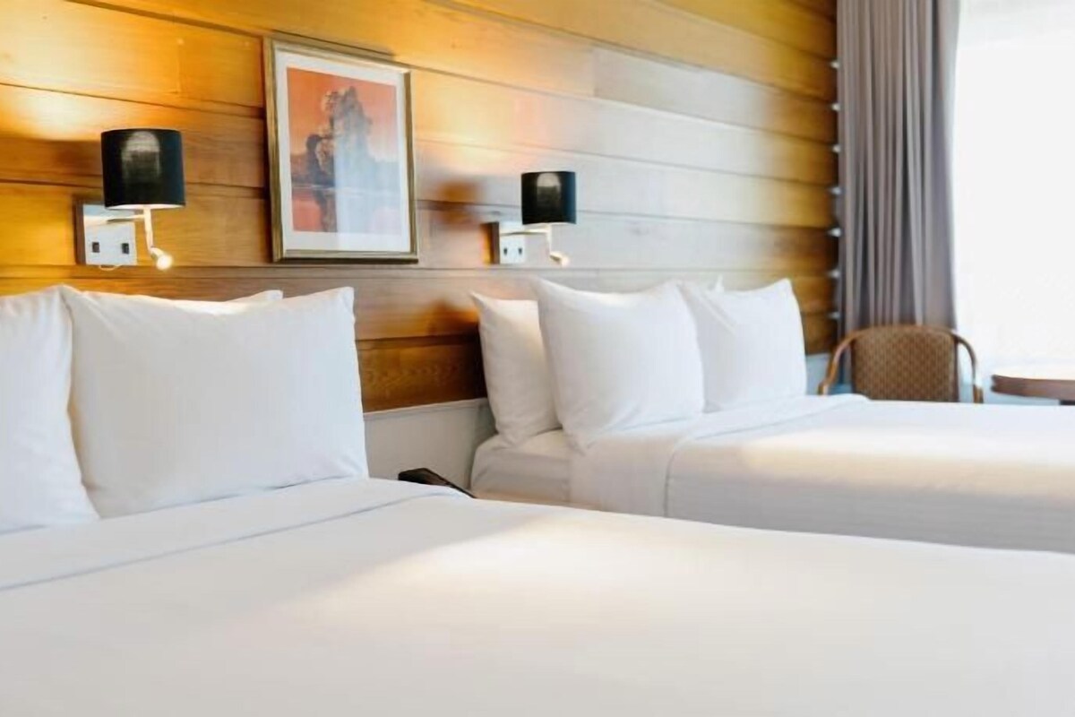 Habitación de hotel con dos camas, cabecero de madera, lámparas de pared y un cuadro enmarcado.