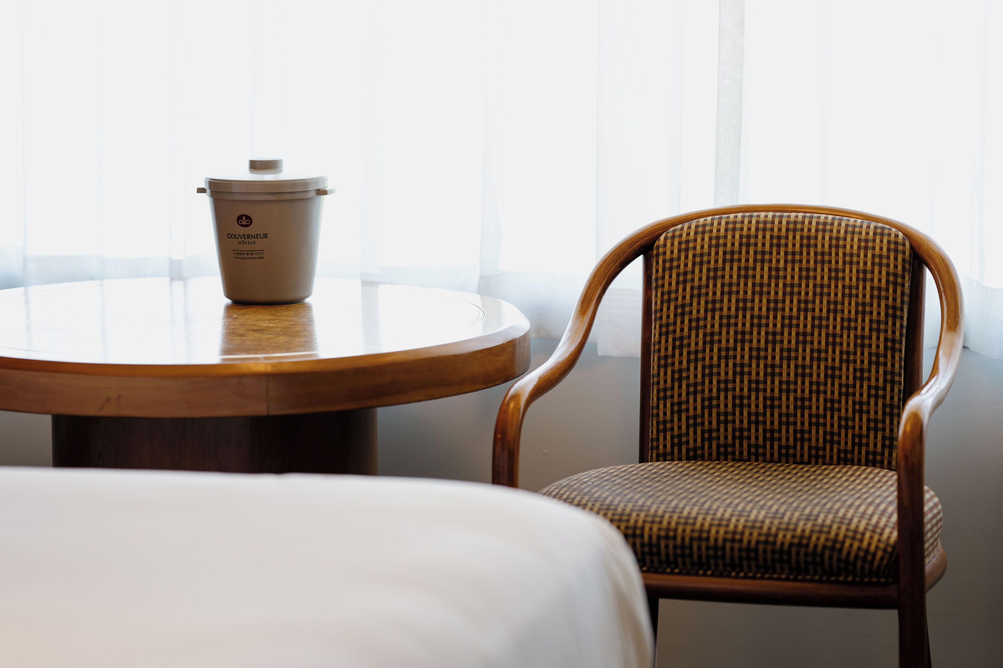 A hotel room with a round wooden table, a wicker chair, and a white bed.