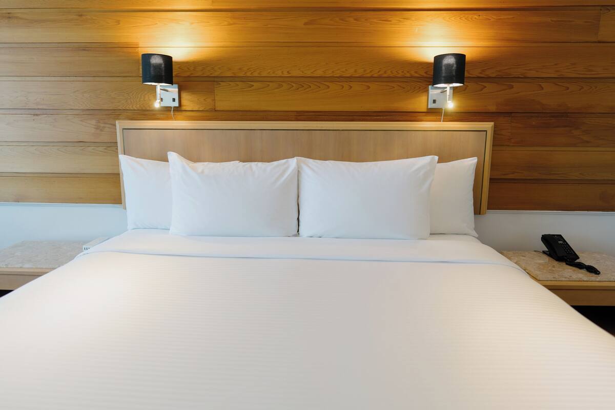 A neatly made bed with white linens, two wall-mounted light fixtures, and a bedside phone on a wooden nightstand.