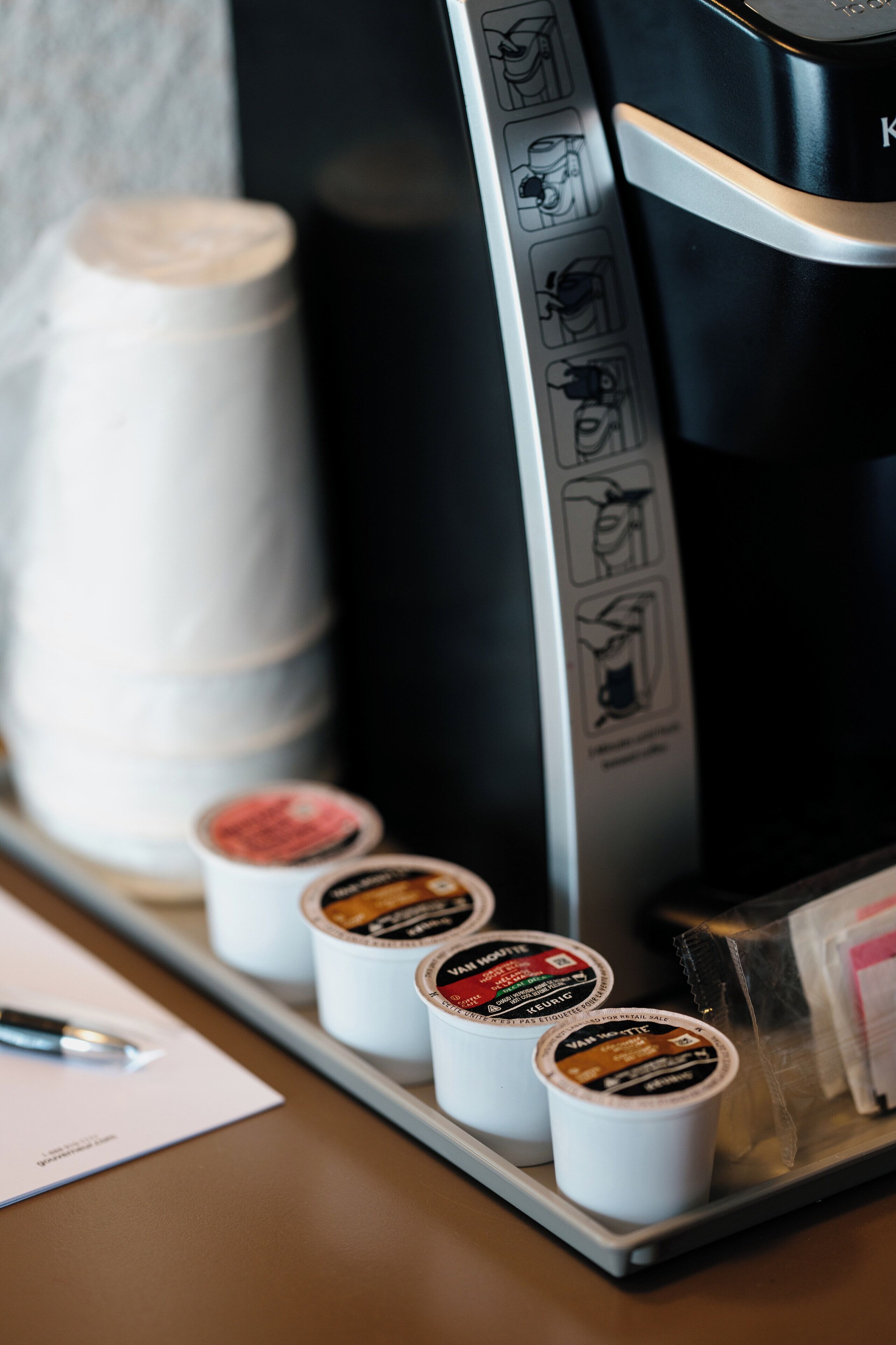 A coffee machine with instructions, a stack of paper cups, and several coffee pods on a tray.