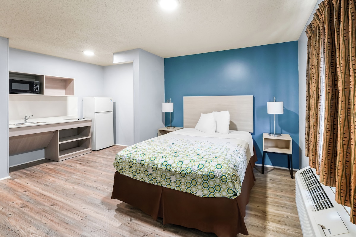 A hotel room with a bed, a desk, a microwave, a refrigerator, a bedside lamp, and a heater.