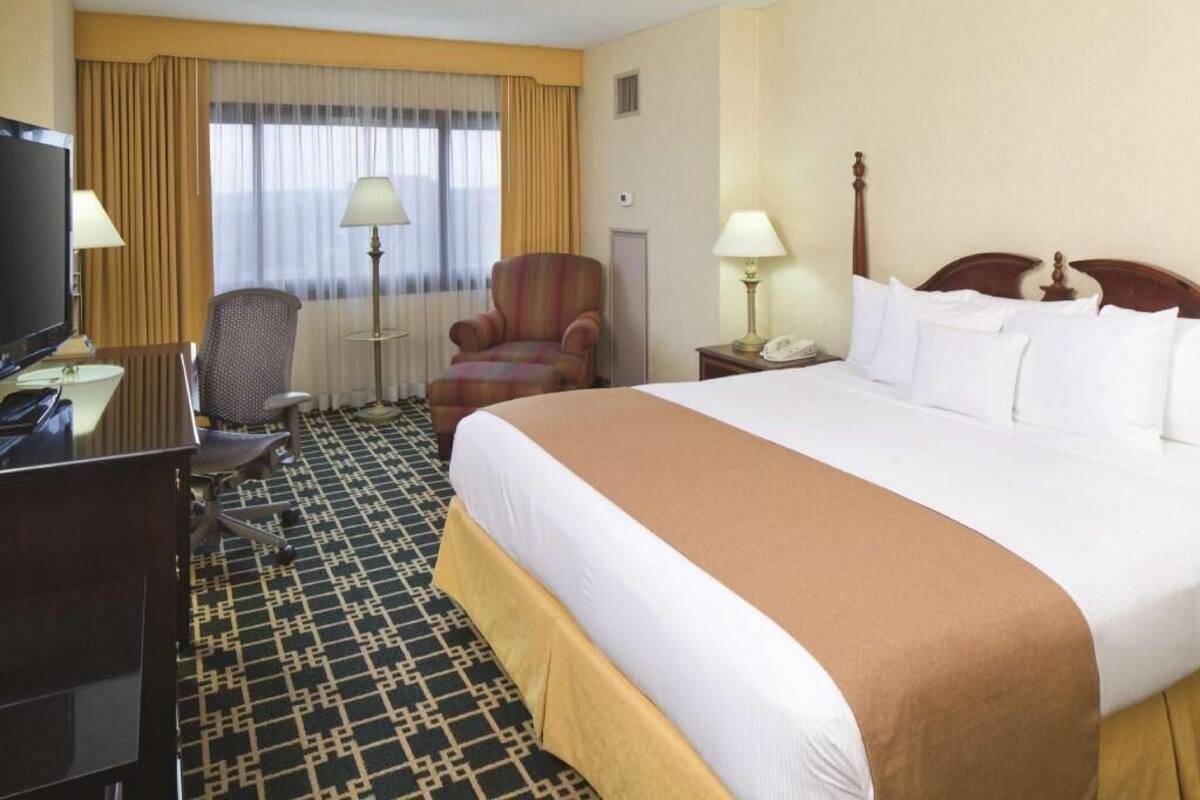 A hotel room with a large bed, a television, a desk with a computer, a chair, and a window with curtains.