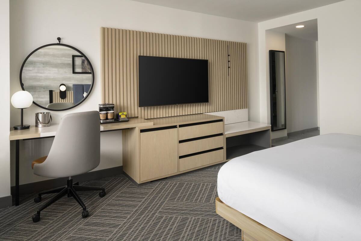 A modern hotel room with a large bed, a desk with a computer, a flat-screen TV, a round mirror, and a wooden panel wall.