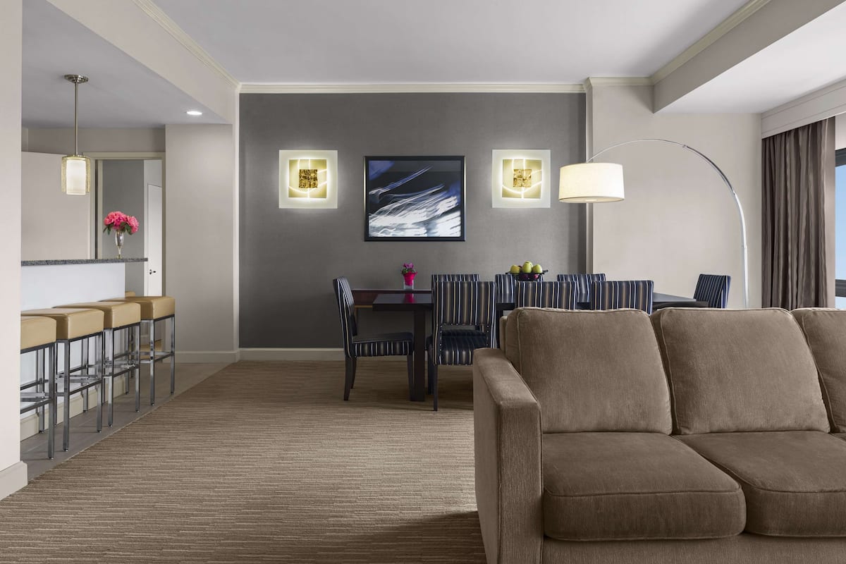 A hotel room with a sofa, a dining table with chairs, a bar area, and a large window with curtains.