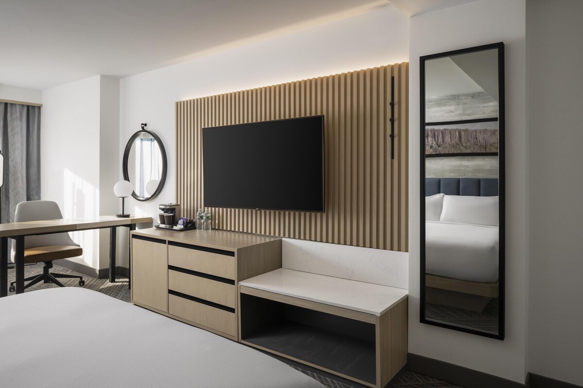A modern hotel room with a flat-screen TV mounted on a wooden panel wall, a large mirror, a bed with a headboard, and a desk with a chair.