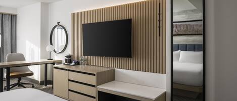 A modern hotel room with a flat-screen TV mounted on a wooden panel wall, a large mirror, a bed with a headboard, and a desk with a chair.