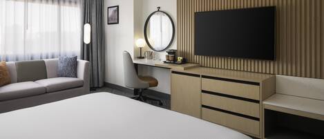 A hotel room with a large bed, a desk with a computer, a TV, and a seating area with a sofa and armchair.