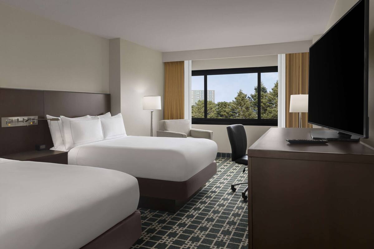 A hotel room with two beds, a desk, a TV, and a window with a city view.