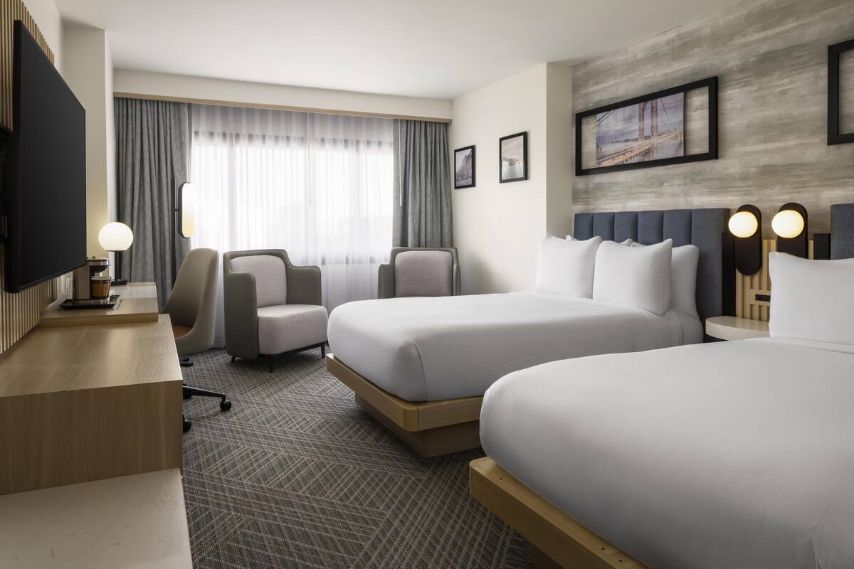 A hotel room with two beds, a desk, a chair, a TV, and a large window with curtains.