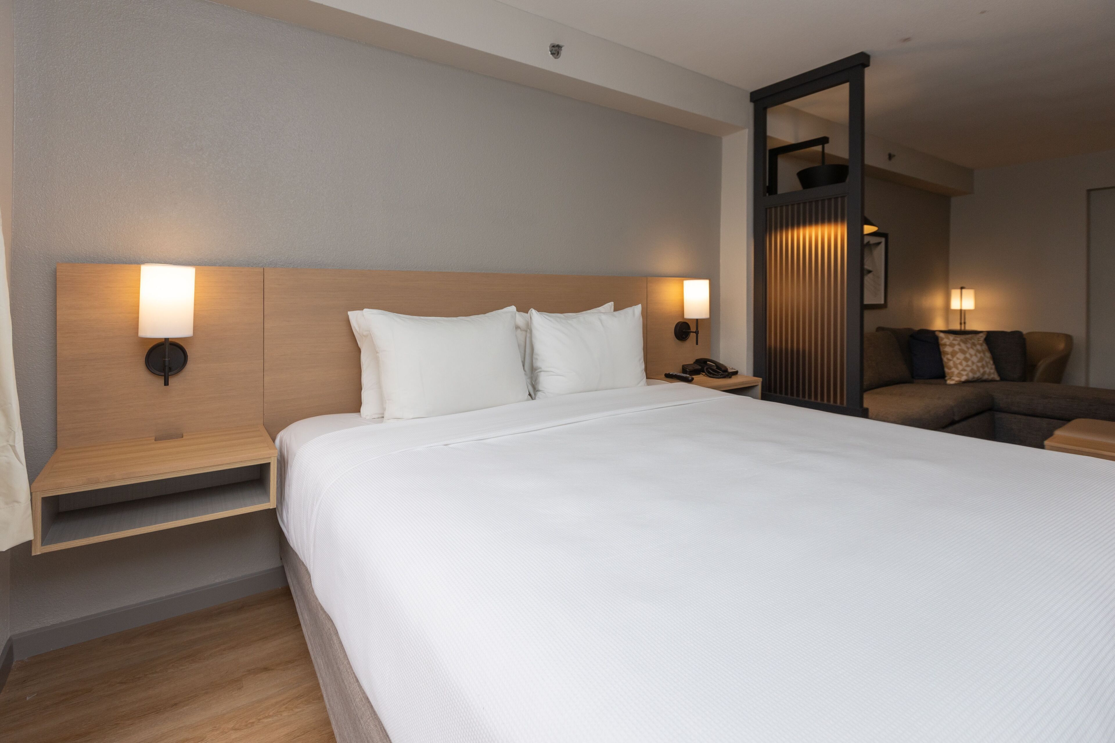 Una camera d'albergo moderna con un letto grande, due lampade da comodino, una testiera in legno, un comodino e un'area salotto con divano e cuscini.