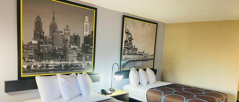 Ein Hotelzimmer mit zwei Betten, jedes mit einer gemusterten Tagesdecke und weißen Kissen. Darüber hängt ein gerahmtes Stadtbild.