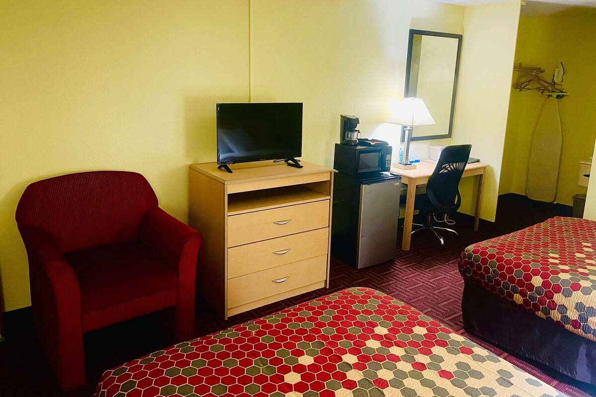 Une chambre d’hôtel avec un téléviseur posé sur un meuble, un bureau avec un ordinateur et une lampe, une chaise rouge, deux lits avec une literie à motifs et un four à micro-ondes.