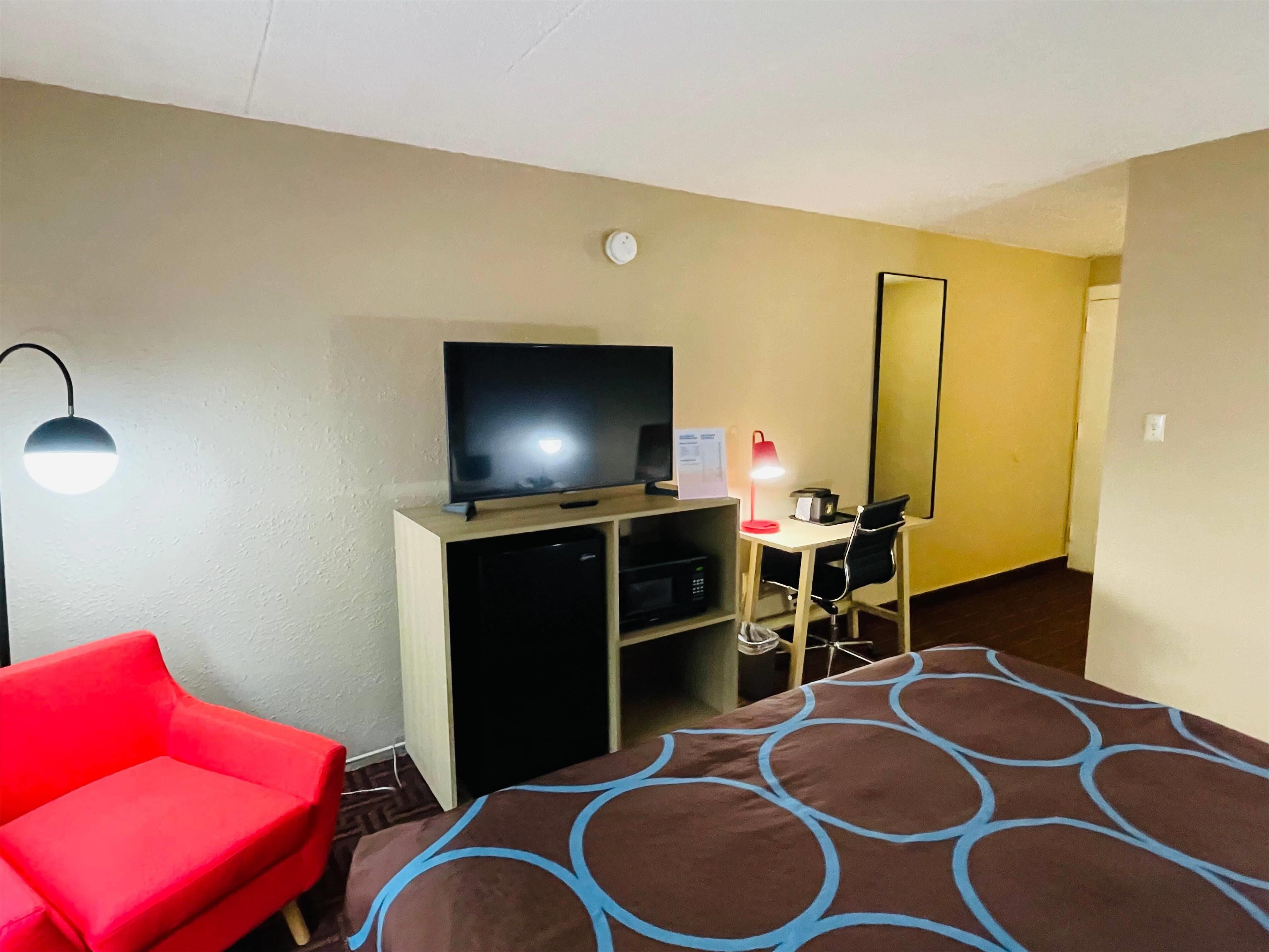 A hotel room with a bed, a desk with a chair, a television, a red chair, and a lamp.